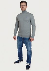 Grauer Rollkragenpullover mit strukturierter Oberfläche, blauem Akzent-Logo, kombiniert mit blauen Jeans und navyblauen Freizeitschuhen.