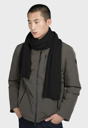 Bufanda negra de canalé hecha de tela suave, colocada de manera casual alrededor del cuello. Se lleva con una chaqueta oscura verde con cremallera. Diseño minimalista.
