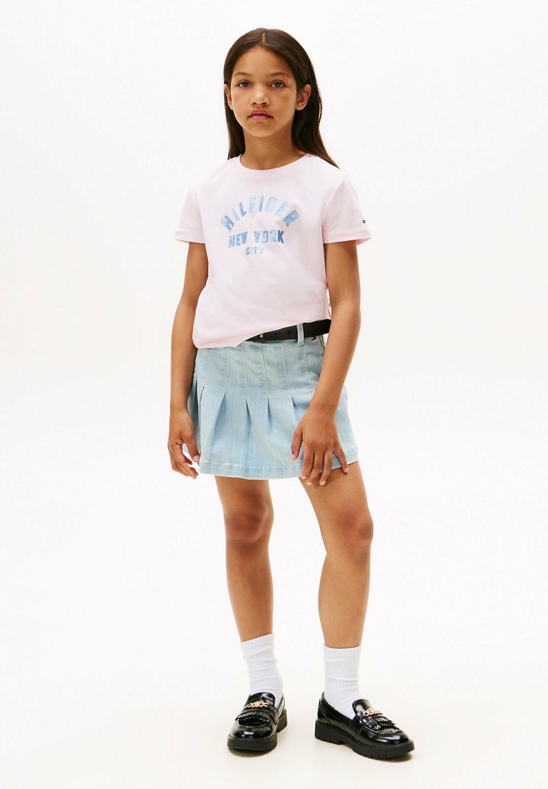 Camiseta de manga corta rosa con impresión azul "Tommy Hilfiger New York City", combinada con una falda plisada azul clara y mocasines negros.