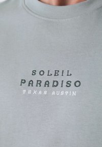 Šviesiai pilkas trumpomis rankovėmis marškinėliai su banguota tekstūra. Ant jų užrašyta "SOLEIL PARADISO" ir "TEXAS AUSTIN" skirtingais juodos ir baltos atspalviais.