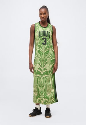 Kvinde stående iført en ærmeløs, grøn, lang Adidas-kjole med abstrakte mønstre, sorte sneakers og hvide sokker mod en ensfarvet baggrund.