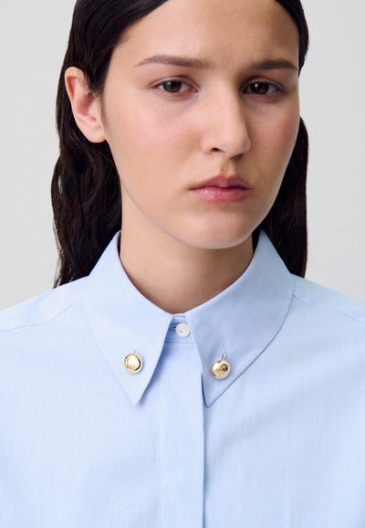 Une femme aux cheveux noirs porte une chemise bleu clair à boutons avec des épingles de col rond dorées sur un fond clair uni.