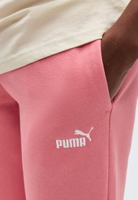 Χέρι στην τσέπη ροζ φόρμας με λευκό λογότυπο Puma, σε συνδυασμό με μπεζ πουκάμισο.