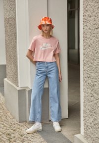 Różowa krótka koszulka Nike z kwiatowym logo, jasnoniebieskie jeansy z szerokimi nogawkami oraz pomarańczowa wiązana kapelusz typu bucket, zestawione z białymi trampkami.