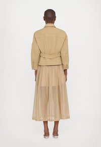 Beige takki, jossa on istuva vyötärö ja selkäkoristelu, yhdistettynä ilmavaan, läpikuultavaan vaaleanbeigeen hameeseen. Kapeat, kärkitossut viimeistelevät asun.