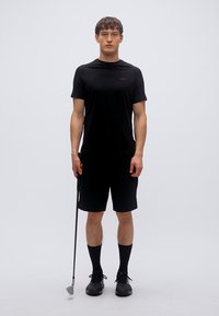 Μαύρο αθλητικό μπλουζάκι και σορτς από αναπνεύσιμο ύφασμα, σε συνδυασμό με μαύρες κάλτσες μέχρι το γόνατο και μαύρα παπούτσια. Κρατάει ένα γκολφ ρόπαλο.