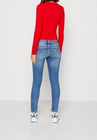 Top rojo de manga larga, jeans ajustados azules. Los jeans cuentan con bolsillos traseros y un parche de cuero en la cintura. Zapatillas blancas con acentos de color.