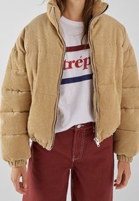 Chaqueta acolchada de pana beige con cuello alto, diseño acolchado y cremallera frontal. Combinada con una camiseta blanca y pantalones burdeos.