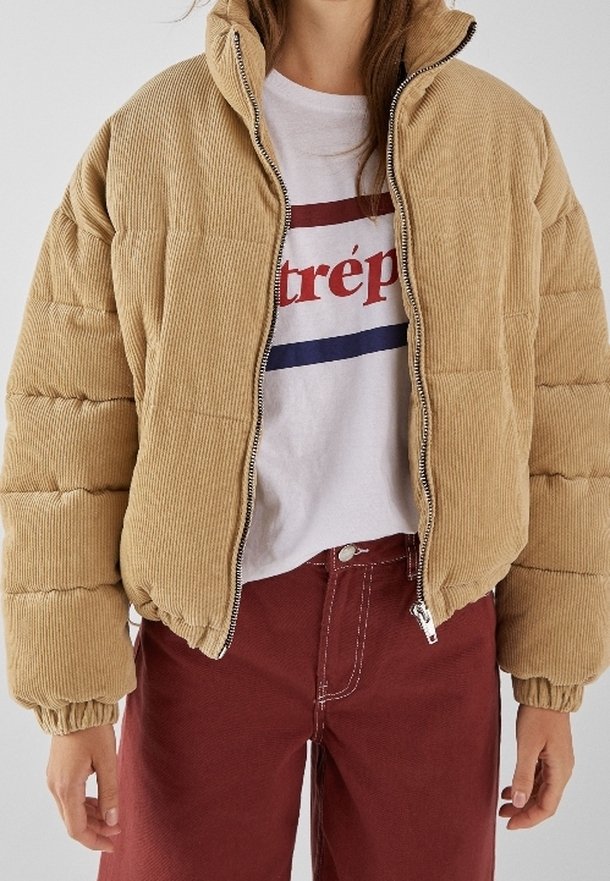 Chaqueta acolchada de pana beige con cuello alto, diseño acolchado y cremallera frontal. Combinada con una camiseta blanca y pantalones burdeos.