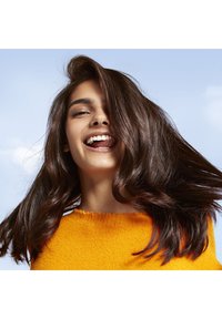 Donna con lunghi capelli castano scuro fluenti, che indossa un maglione arancione brillante e texture, sorridente su uno sfondo di cielo azzurro.