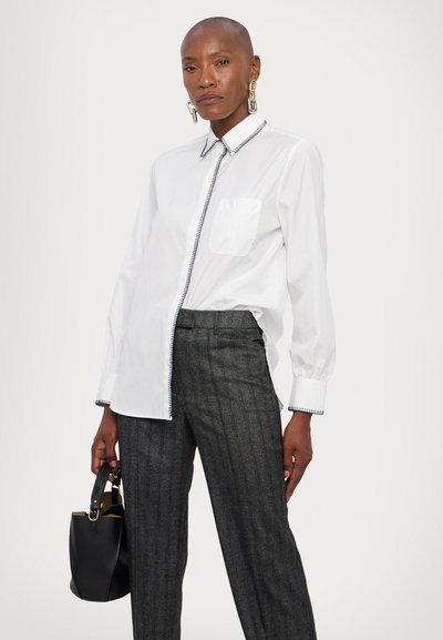 White long-sleeve shirt with a patch pocket and contrasting blue trim, paired with gray pinstriped trousers. Black handbag held in hand.