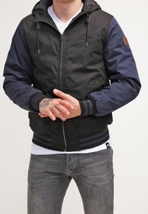 Mann trägt schwarze und marineblaue Kapuzenjacke mit gestreiften Bündchen, weißes T-Shirt, graue Jeans und hat tätowierte Hände, die vor ihm verschränkt sind.