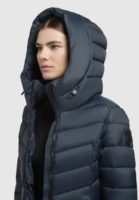 Sötétkék púderdzseki magas gallérral és nagy kapucnival, steppelt részekkel és cipzáras záródással. Logós jelvény az ujjon.