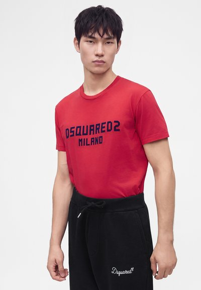 Jeune homme portant un t-shirt rouge Dsquared2 Milano et un pantalon noir avec une taille à cordon, debout devant un fond clair uni.