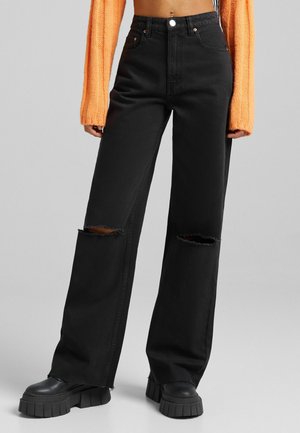 Jeans noirs taille haute avec des jambes larges, présentant des déchirures aux genoux. Fabriqués en denim, ils sont associés à des chaussures à plateforme noires épaisses.