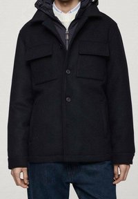 Černá bunda na knoflíky s kapucí, vybavená dvěma velkými kapsami na hrudi a texturovaným materiálem. Nositelná přes šedý vrstvený top a modré džíny.