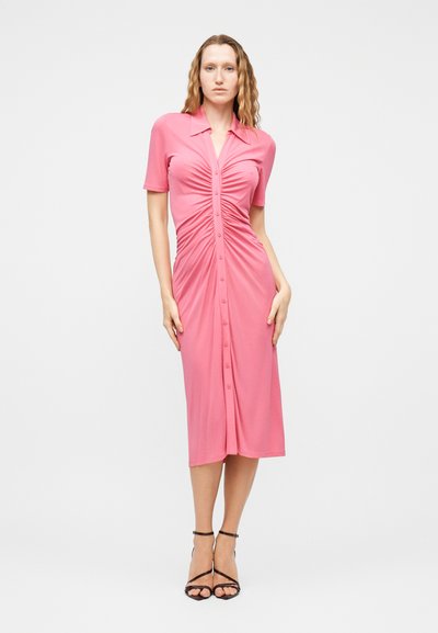 Γυναίκα που φοράει ροζ ρουστίκ φόρεμα μεσαίου μήκους με κουμπιά και κοντά μανίκια, μαζί με μαύρα ψηλοτάκουνα σανδάλια με λουράκια, στέκεται μπροστά σε απλό φόντο.