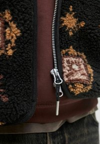 Giacca di pile nera con motivi astratti marroni, cerniera argentata e rifiniture nere. Indossata sopra una maglietta bordeaux e jeans scuri.