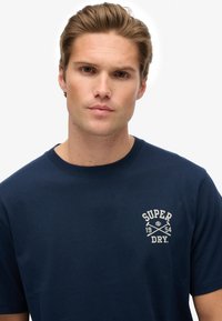 Joven con cabello castaño claro que lleva una camiseta azul marino con el logo blanco "Super Dry 1954" en el pecho, contra un fondo blanco.