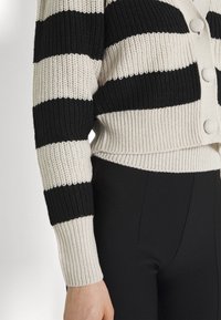 Cárdigan a rayas en negro y crema con textura acanalada, que presenta botones grandes y un diseño corto, combinado con pantalones ajustados negros.