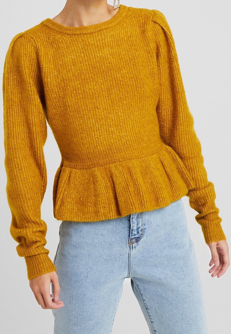 Pull côtelé jaune moutarde à manches longues avec épaules bouffantes et bas péplum, porté avec un jean taille haute bleu clair.