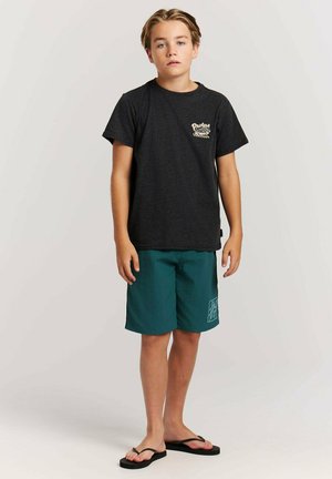 Jongen met blond haar, gekleed in een zwart grafisch T-shirt, teal knielange shorts en zwarte slippers, staand tegen een effen achtergrond.