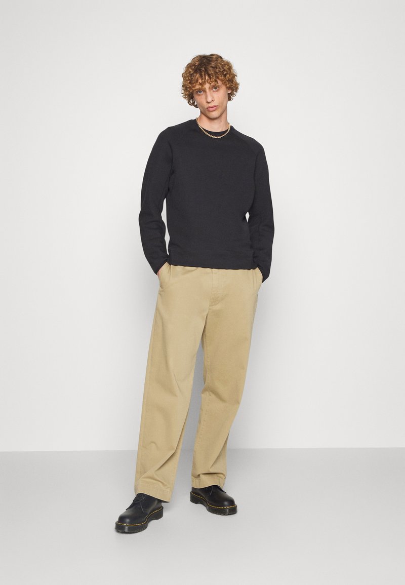 Sweatshirt noir à col rond, pantalons larges beiges, et chaussures noires avec des coutures jaunes. La tenue présente une coupe décontractée et un design simple.