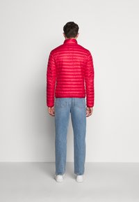 Červená péřová bunda s vysokým límcem, která má prošívaný design. V kombinaci s modrými džíny a bílými teniskami, pohled zezadu.