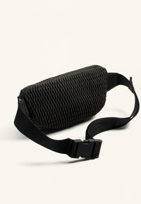 Geantă crossbody țesută de culoare neagră, cu o suprafață texturată, dotată cu o curea ajustabilă rezistentă și o închidere cu cataramă din plastic. Formă compactă, rectangulară.