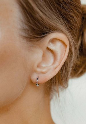 Silberne Creolenohrringe mit mehrfarbigen Edelsteindetails, die eine glatte, reflektierende Oberfläche und ein gewundenes Design aufweisen. Am Ohr getragen.