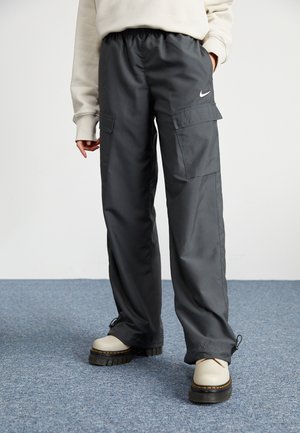 Personne portant un pantalon cargo ample gris foncé avec des poches latérales et des chaussures blanches, debout sur un sol recouvert de moquette bleue.