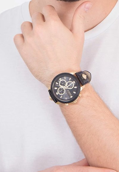 Čierne chronografické hodinky so zlatými akcentmi, veľkým okrúhlym ciferníkom a koženým remienkom, predvádzané na zápästí s pozadím svetlej košele.