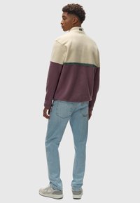 Sudadera de forro polar con bloques de color en crema, púrpura y verde azulado. Presenta un cuello alto y una superficie texturada. Se lleva con jeans de color azul claro y zapatillas.