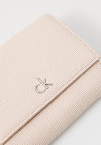 Gaiši rozā teksturēta maka, kas izgatavots no sintētiska materiāla, ar sudraba logotipa akcentu un minimālistisku dizainu ar redzamām šuvēm.