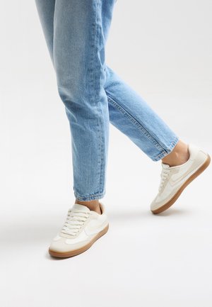 Sneaker bianca con tomaia liscia, suola in gomma e design con lacci. Accentata da un logo sottile e una suola marrone chiaro, abbinata a jeans blu.