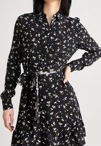 Černé květinové šaty s bílými a žlutými květinovými vzory. Mají zapínání na knoflíky vpředu, dlouhé rukávy a pásek s textem "Tommy Jeans".