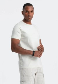 Camiseta de algodón beige claro con cuello redondo, mangas cortas y sutiles acentos de costura. El modelo lleva una pulsera y pantalones cargo.