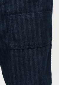 Κοντινή λήψη παντελονιού με ύφασμα σε σχέδιο ψαροκόκκαλο σε σκούρο μπλε χρώμα, με ορατή ορθογώνια τσέπη στο πλάι.