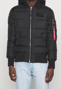 Sort quilted hættejakke med frontlynlås og rød "Remove Before Flight"-tag på venstre arm, båret med lyseblå jeans.