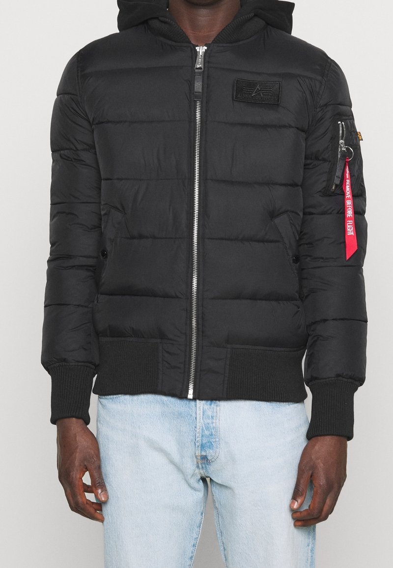 Sort quilted hættejakke med frontlynlås og rød "Remove Before Flight"-tag på venstre arm, båret med lyseblå jeans.