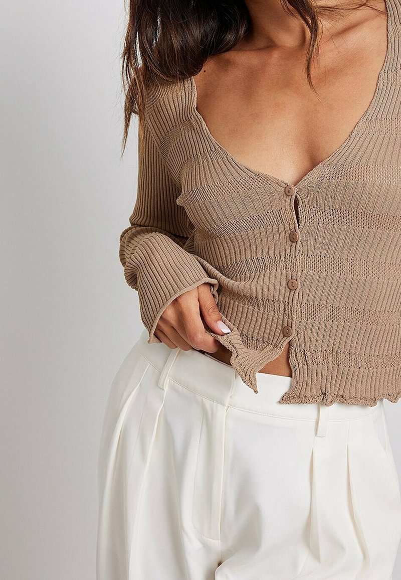 En kvinna modellerar en ribbad, beige cardigan med djup V-ringning och knappar, i kombination med högmidjade vita byxor, vilket visar en stilig look.