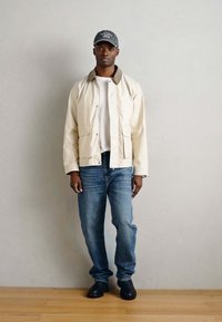 Ανοιχτό μπεζ, αδιάβροχο μπουφάν με φερμουάρ, δύο τσέπες και καφέ γιακά, φορεμένο πάνω από λευκό πουκάμισο και ανοιχτό μπλε τζιν. Μαύρα παπούτσια.