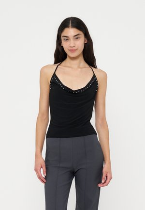 Femme aux longs cheveux foncés portant un haut noir dos-nu avec de petits clous et un pantalon taille haute gris, se tenant devant un fond uni.
