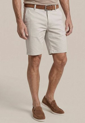 Man met beige korte broek, een bruine leren riem en bruine instappers staat met ontspannen handen langs het lichaam.