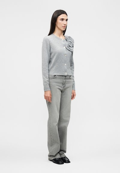 Γυναίκα στέκεται φορώντας ανοιχτό γκρι ζακέτα με κουμπιά και λουλουδένια λεπτομέρεια, γκρι τζιν και μαύρα παπούτσια σε λευκό φόντο.