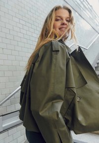 Olijfgroen oversized jack met gestructureerde schouders, knoopdetails en gelaagd ontwerp. Heeft een soepele, licht glanzende textuur.