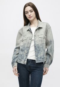 Jauna sieviete ar gariem brūniem matiem, tērpusies izbalējušā džinsa jakā, baltā krekliņā un tumši zilās džinsās, balta fona priekšā.