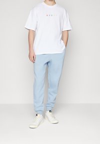 Bílá bavlněná tričko s vícero barevným textem, doplněné světle modrými fleecovými tepláky. Model má na sobě bílé tenisky s krémovými akcenty.