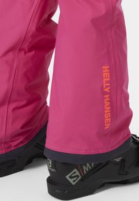 Růžové voděodolné lyžařské kalhoty s hladkým povrchem, zdobené logem Helly Hansen v oranžové barvě, doplněné černými lyžařskými botami s strukturovanou podrážkou.