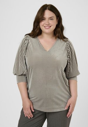 Mujer sonriente con cabello castaño que lleva una blusa plateada brillante de rayas con escote en V y mangas abullonadas de 3/4, vestida con pantalones gris oscuro, de pie frente a un fondo blanco.